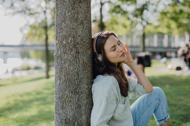 Porträt einer jungen Frau, die im Stadtpark über Kopfhörer Musik hört. - HPIF12499