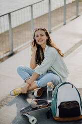 Happy young woman sitting on a skateboard and calling at city bridge. Youth culture and commuting concept. - HPIF12501