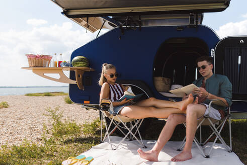 Ein glückliches junges Paar sitzt vor einem Van, zeltet und liest ein Buch. - HPIF12553