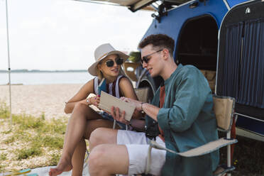 Ein glückliches junges Paar sitzt vor einem Van, zeltet und liest ein Buch. - HPIF12554