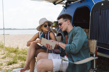 Happy young couple sitting together in front of van, camping and reading a book. - HPIF12554