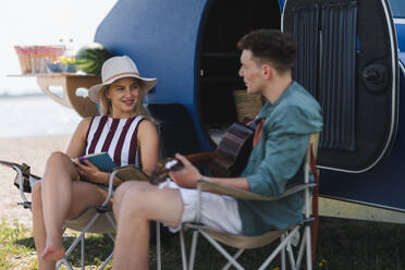 Happy young couple sitting together in front of van, camping and playing at a guitair. - HPIF12556