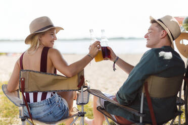 Happy young couple sitting together in front of van, camping and having toast with bottled beer. - HPIF12568