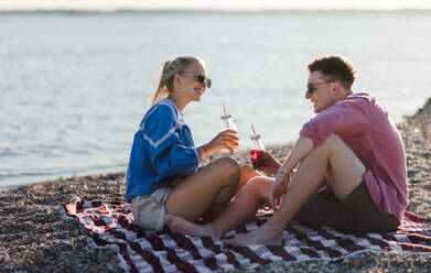 Glückliches junges Paar, das sich am Strand verabredet, auf der Decke sitzt und mit Flaschenbier anstößt und die gemeinsame Urlaubszeit genießt. - HPIF12585
