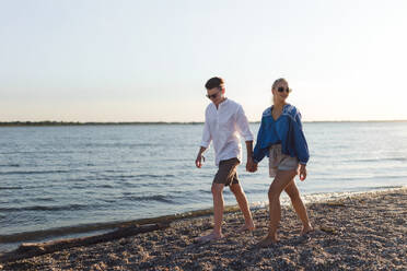 Glückliches junges Paar, das bei Sonnenuntergang am Strand spazieren geht und sich auf ein Picknick vorbereitet. - HPIF12591