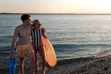 Ein glückliches junges Paar in Badeanzügen posiert mit Schwimmrad am Strand bei Sonnenuntergang. - HPIF12598