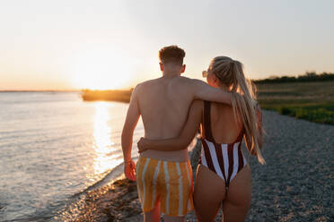 Eine Rückansicht eines jungen Paares in gestreiften Badeanzügen, das bei Sonnenuntergang am Strand spazieren geht. - HPIF12601