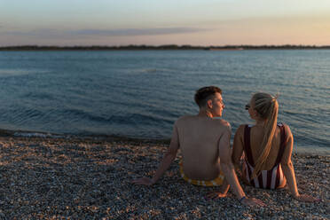 Eine Rückansicht eines jungen Paares in gestreiften Badeanzügen, das bei Sonnenuntergang am Strand sitzt. - HPIF12604