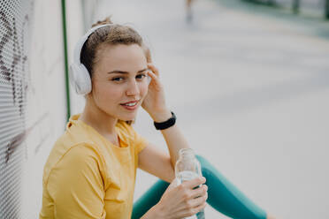 Junge Frau trinkt Wasser und hört Musik über Kopfhörer, beim Joggen in der Stadt, gesunder Lebensstil und Sportkonzept. - HPIF12611