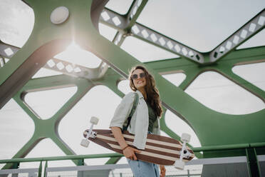 Junge Frau, die mit einem Skateboard auf einer Brücke in der Stadt spazieren geht, Jugendkultur und Pendeln. - HPIF12620