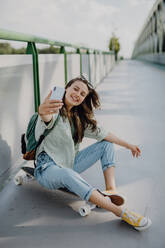 Eine fröhliche junge Frau macht ein Selfie, während sie auf einem Skateboard an einer Stadtbrücke sitzt und die Jugendkultur und das Pendeln repräsentiert - HPIF12621