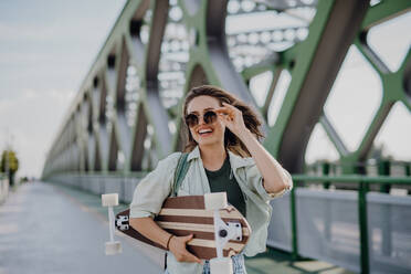 Young woman walking on city bridge with a skateboard. Youth culture and commuting concept. - HPIF12622