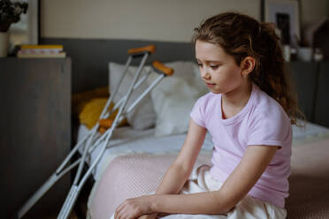 Kleines unglückliches Mädchen mit gebrochenem Bein, das auf einem Bett in ihrem Zimmer sitzt. - HPIF12678