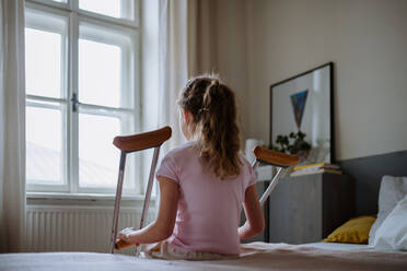 Rear view of little girl with broken leg sitting on a bed in her room. - HPIF12679