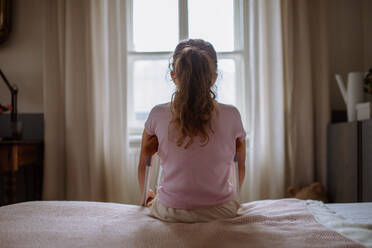 Rear view of little unhappy girl with broken leg sitting on a bed in her room. - HPIF12680