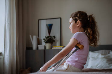 Little unhappy girl with broken leg sitting on a bed in her room. - HPIF12681