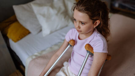 Little unhappy girl with broken leg sitting on a bed in her room. - HPIF12682