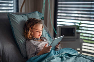 Gelangweiltes kleines Mädchen, das im Bett im Krankenhaus liegt und ein Tablet benutzt. - HPIF13065
