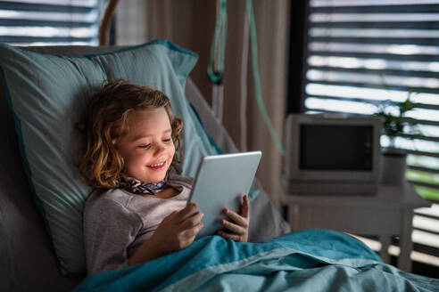 Gelangweiltes kleines Mädchen, das im Bett im Krankenhaus liegt und ein Tablet benutzt. - HPIF13099