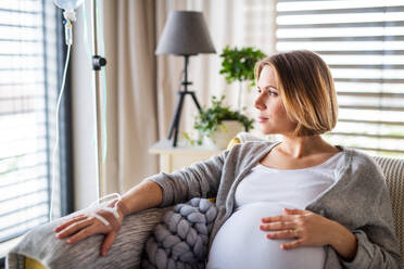 A portrait of pregnant woman with IV drip indoors at home or in hospital, sitting on sofa. - HPIF13138