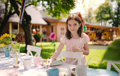 Porträt eines kleinen Mädchens, das an einem Tisch im Garten im Sommer auf einer Party steht und in die Kamera schaut. - HPIF13180