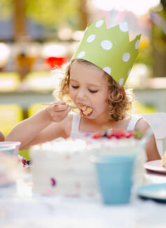 Kleines Mädchen mit Partyhut sitzt im Sommer draußen im Garten und isst Kuchen - ein Konzept für eine Feier. - HPIF13226