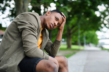 Trauriger und frustrierter junger schwarzer Mann sitzt auf einer Bank im Freien in der Stadt, Black Lives Matter Konzept. - HPIF13257
