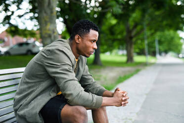 Trauriger und frustrierter junger schwarzer Mann sitzt auf einer Bank im Freien in der Stadt, Black Lives Matter Konzept. - HPIF13258