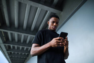 Niedriger Blickwinkel eines jungen schwarzen Mannes im Freien in der Stadt, der ein Smartphone benutzt. - HPIF13283