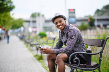 Cheerful young black man commuter sitting on bench outdoors in city, laughing. - HPIF13290