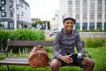 Cheerful young black man commuter sitting on bench outdoors in city, resting. - HPIF13294