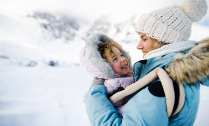 Portrait of mother with happy small daughter in carrier standing in winter nature, resting. - HPIF13328