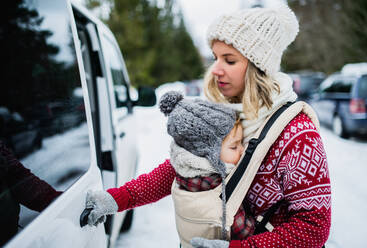 Portrait of mother with sleeping small daughter in carrier standing by car in winter nature. - HPIF13373