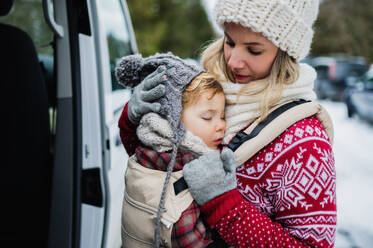 Portrait of mother with sleeping small daughter in carrier standing by car in winter nature. - HPIF13374