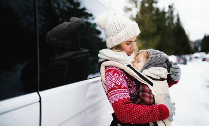 Portrait of mother with sleeping small daughter in carrier standing by car in winter nature. - HPIF13375