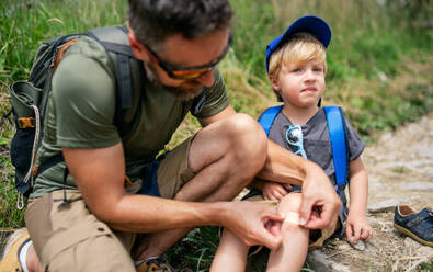 Älterer Vater mit kleinem Sohn beim Wandern in der sommerlichen Natur, Pflaster auf das Knie kleben. - HPIF13389