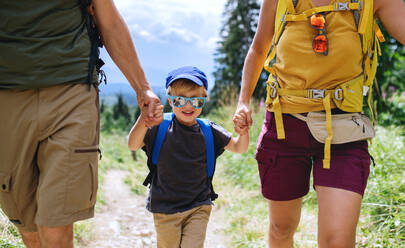 Vorderansicht eines kleinen Jungen mit nicht erkennbaren Eltern, die im Sommer in der Natur wandern und sich an den Händen halten. - HPIF13393