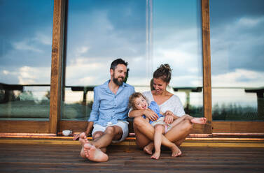 Glückliche Familie mit kleiner Tochter sitzt auf der Veranda einer Holzhütte, Urlaub in der Natur Konzept. - HPIF13472