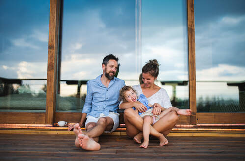 Happy family with small daughter sitting on patio of wooden cabin, holiday in nature concept. - HPIF13472