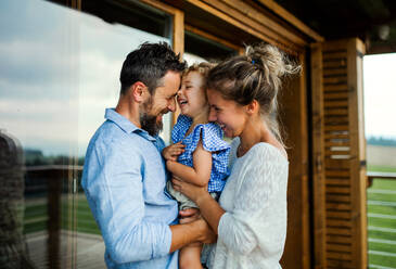 Happy family with small daughter standing on patio of wooden cabin, holiday in nature concept. - HPIF13473