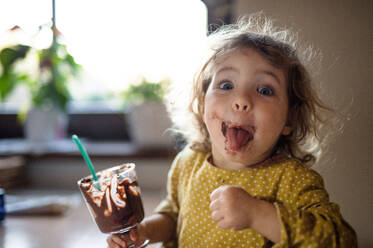 Glückliches kleines Mädchen mit schmutzigem Mund drinnen in der Küche zu Hause, das Pudding isst. - HPIF13490