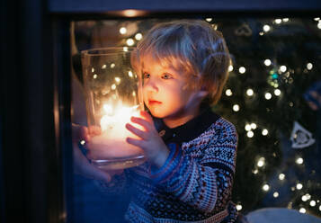 Porträt eines glücklichen kleinen Jungen, der zu Hause zu Weihnachten eine Kerze in der Hand hält. - HPIF13551