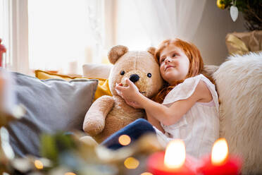 Glückliches kleines Mädchen, das zu Weihnachten zu Hause auf dem Sofa sitzt und einen Teddybären umarmt. - HPIF13585
