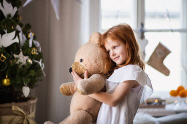 Porträt eines kleinen Mädchens, das zu Weihnachten zu Hause steht und einen Teddybären umarmt. - HPIF13587