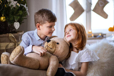 Small girl and boy sitting on sofa indoors at Christmas, holding big teddy bear. - HPIF13589