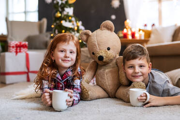 Kleines Mädchen und Junge im Schlafanzug auf dem Fußboden zu Hause an Weihnachten, Blick in die Kamera. - HPIF13608