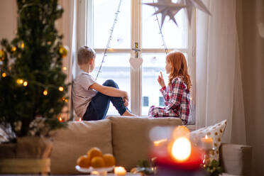 Kleines Mädchen und Junge im Schlafanzug zu Hause an Weihnachten, sitzen auf der Fensterbank und unterhalten sich. - HPIF13609