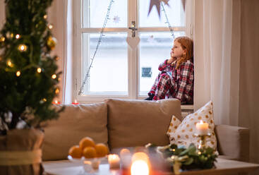 Seitenansicht des Porträts eines kleinen traurigen Mädchens im Schlafanzug zu Hause zu Weihnachten, das am Fenster sitzt. - HPIF13611