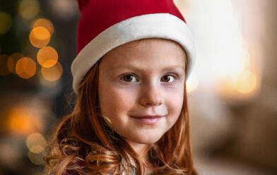 Nahaufnahme eines kleinen Mädchens mit Weihnachtsmannmütze zu Hause an Weihnachten, das in die Kamera schaut. - HPIF13614