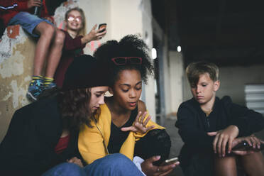 Vorderansicht einer Gruppe Jugendlicher, die in einem verlassenen Gebäude sitzen und Smartphones benutzen. - HPIF13627